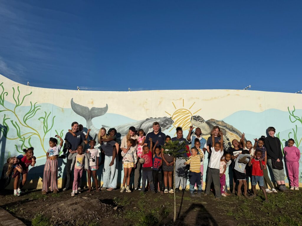 Children, volunteers and staff celebrating a tree planting in front of the KuCoNa whale mural