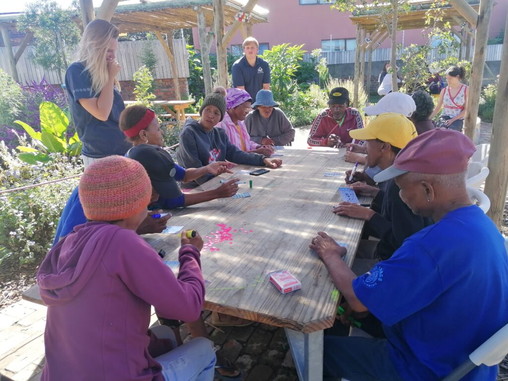 Community members playing bingo at a long outdoor table in the KuCoNa garden